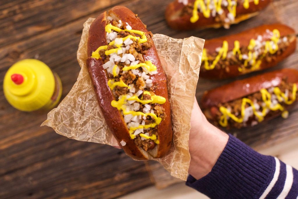 A hand holds out a tangy, juicy-looking chili dog that’s wrapped in a fresh-steamed bun